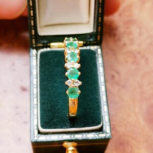 May include: A gold ring featuring a row of alternating round green emeralds and clear white diamonds set in a vertical line. The ring is displayed upright inside a dark green velvet-lined jewelry box with a hinged lid and gold trim.