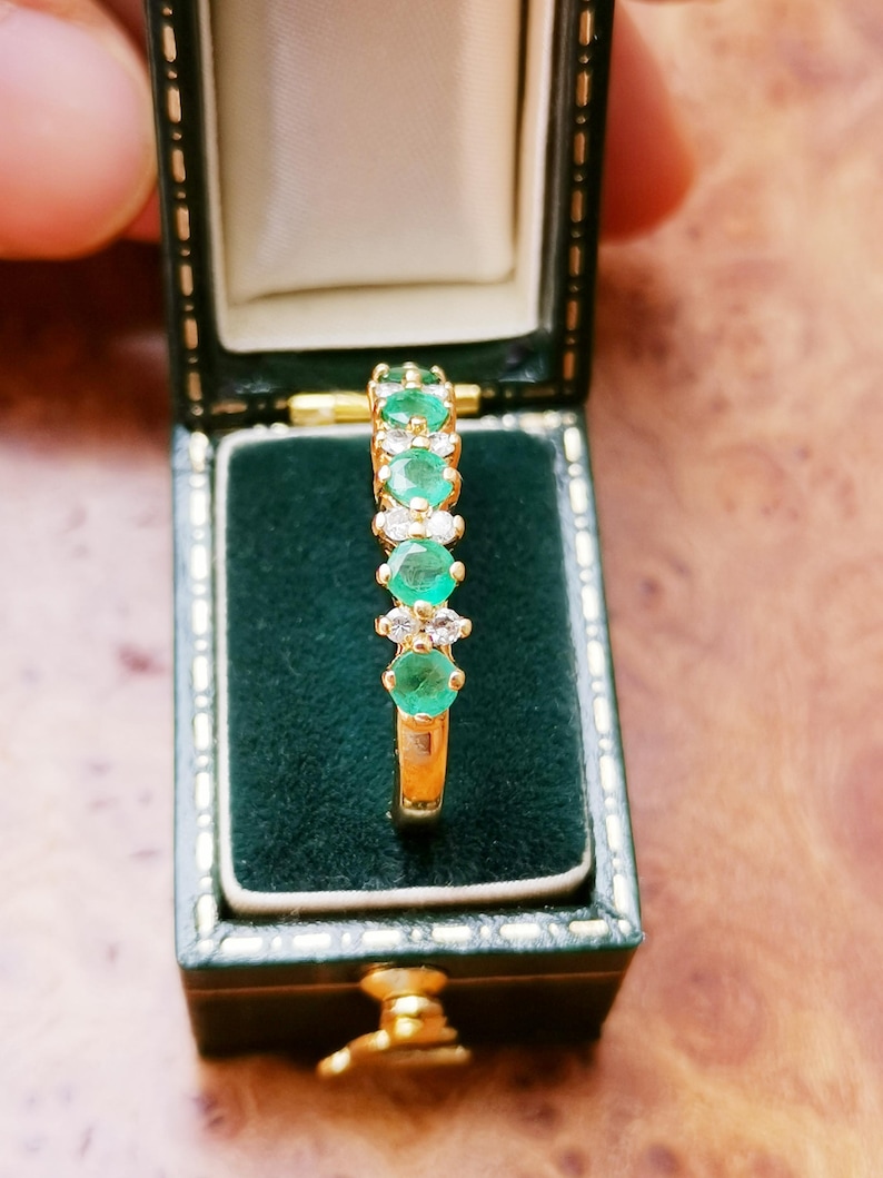 May include: A gold ring featuring a row of alternating round green emeralds and clear white diamonds set in a vertical line. The ring is displayed upright inside a dark green velvet-lined jewelry box with a hinged lid and gold trim.