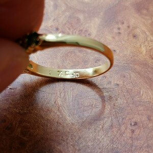 May include: A gold-toned ring featuring a smooth, polished band with the letters XC and the number 10 engraved on the interior surface. The ring has a dark green gemstone setting visible at the top, showcasing a metallic finish and clear markings.
