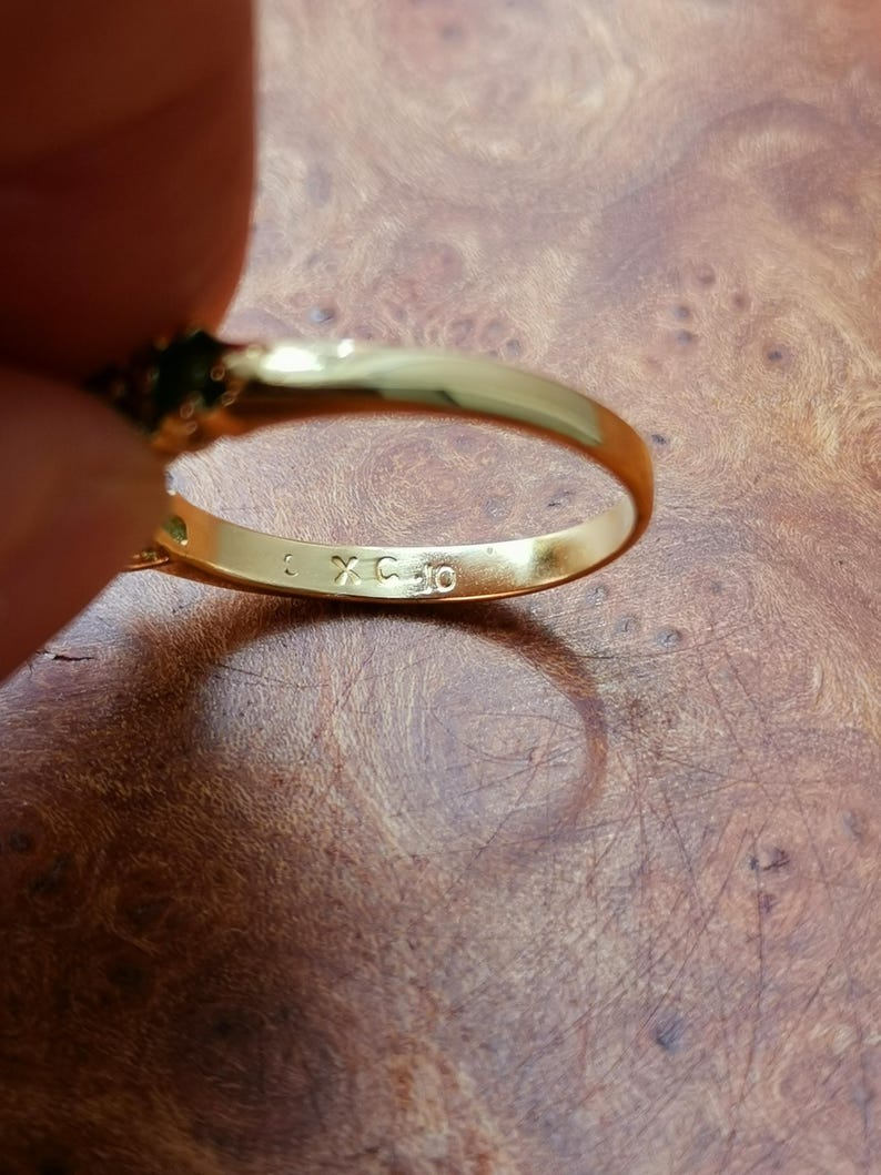 May include: A gold-toned ring featuring a smooth, polished band with the letters XC and the number 10 engraved on the interior surface. The ring has a dark green gemstone setting visible at the top, showcasing a metallic finish and clear markings.