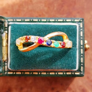 Può includere: Un anello dorato a forma di infinito incastonato con una serie di pietre preziose colorate. L'anello è esposto all'interno di un portagioie di velluto verde con finiture dorate. Le pietre preziose includono pietre rosse, bianche, rosa, blu e verdi.