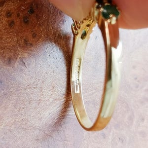 May include: A gold ring featuring a dark green gemstone set at the top. The inner band of the ring displays clear hallmarks, including the text 750, indicating the metal purity. The ring has a polished finish and a smooth, curved metal band.