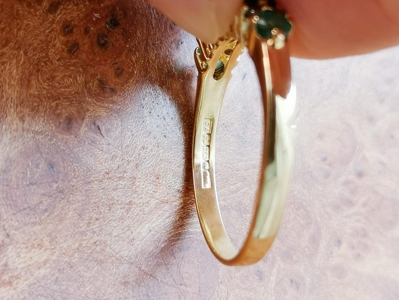 May include: A gold ring featuring a dark green gemstone set at the top. The inner band of the ring displays clear hallmarks, including the text 750, indicating the metal purity. The ring has a polished finish and a smooth, curved metal band.