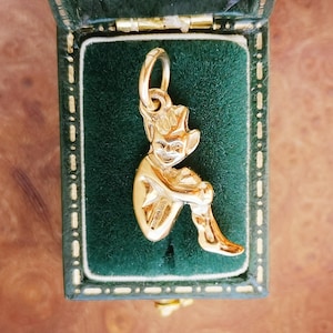 Puede incluir: Un charm de color dorado con forma de elfo o duende sentado, con un gorro puntiagudo y zapatos curvados. El charm se muestra en una caja de joyería de terciopelo verde con ribete dorado. El charm es probablemente de oro.