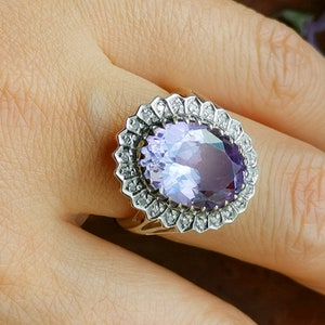 Op de afbeelding: Een ring met een grote ovale amethist-edelsteen omringd door een halo van kleinere diamanten. De ring is gezet in een band van wit metaal.