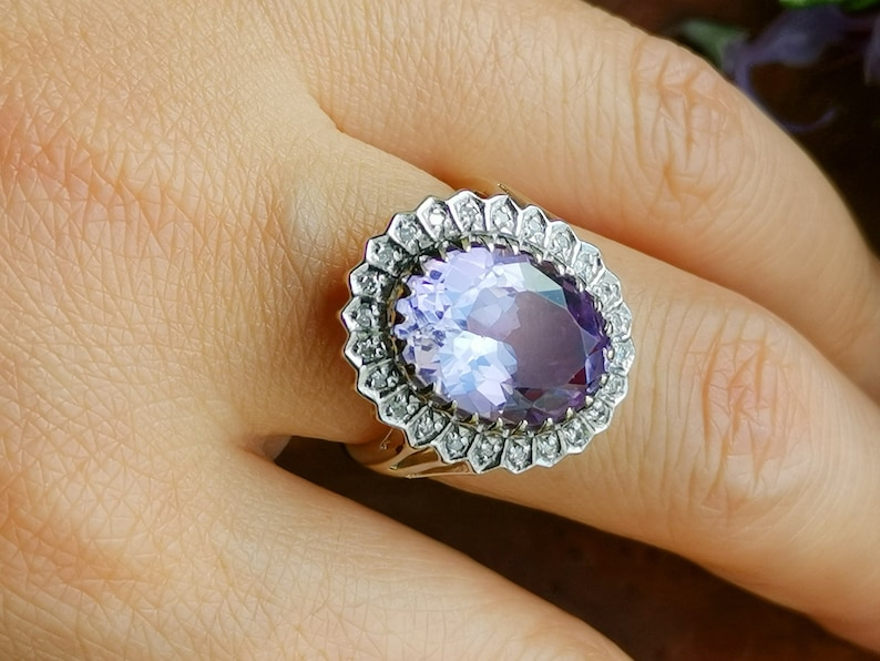 Op de afbeelding: Een ring met een grote ovale amethist-edelsteen omringd door een halo van kleinere diamanten. De ring is gezet in een band van wit metaal.
