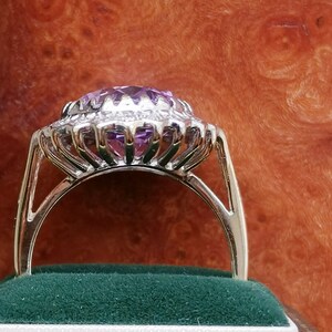 Op de afbeelding: Een zilveren ring met een grote, ovale, paarse edelsteen omringd door een halo van kleinere diamanten. De ring is gezet op een band van wit metaal.