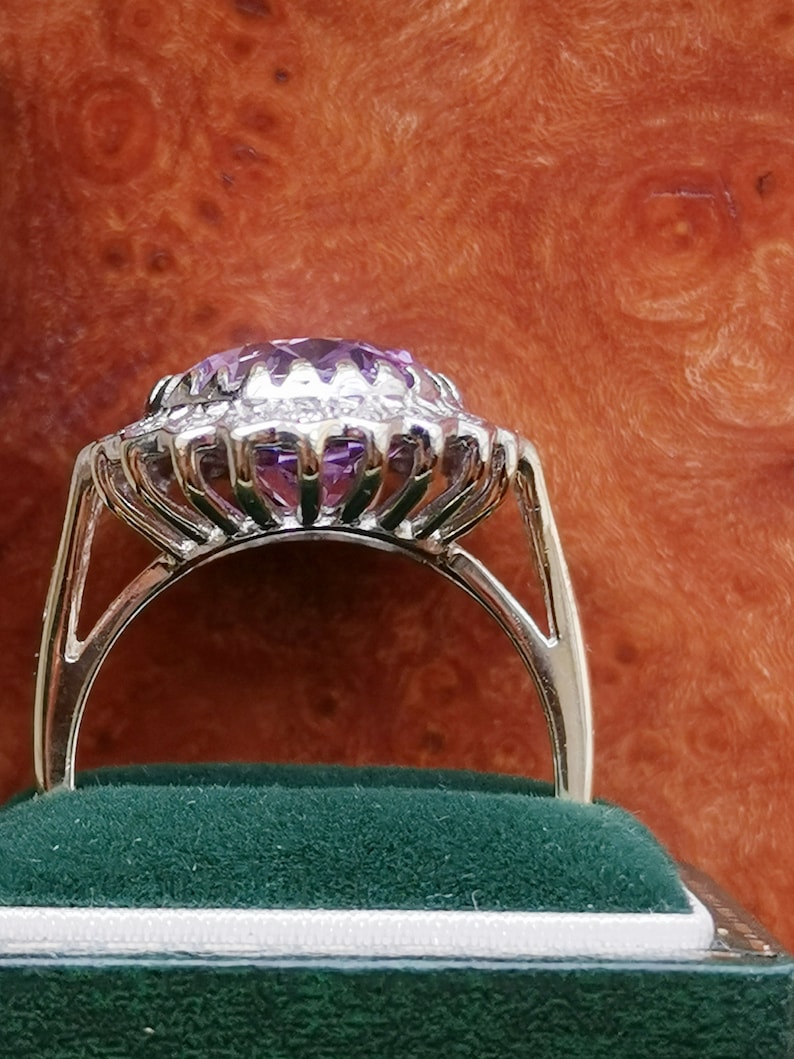 Op de afbeelding: Een zilveren ring met een grote, ovale, paarse edelsteen omringd door een halo van kleinere diamanten. De ring is gezet op een band van wit metaal.