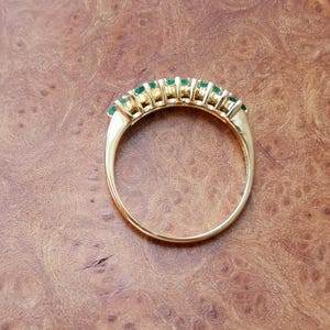 May include: A gold ring featuring a row of seven small, round, vibrant green gemstones set along the top curve of the band. The metal band is polished and smooth, showcasing a classic circular design with the stones arranged in a horizontal line.