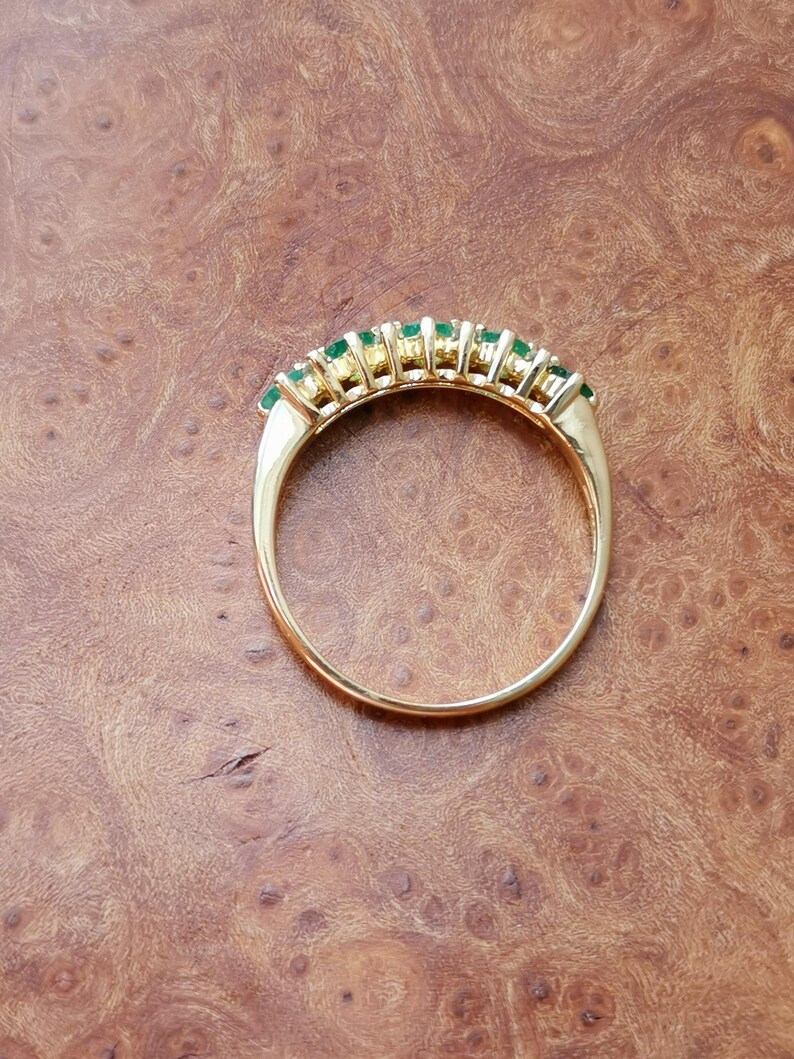 May include: A gold ring featuring a row of seven small, round, vibrant green gemstones set along the top curve of the band. The metal band is polished and smooth, showcasing a classic circular design with the stones arranged in a horizontal line.