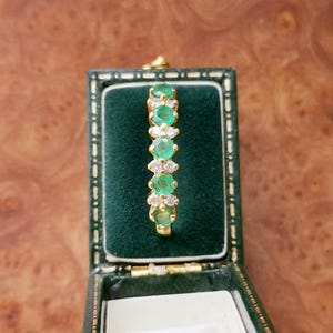 May include: A gold ring featuring a row of five round green emeralds alternating with small clear white diamonds, set in a vertical arrangement within a dark green velvet-lined jewelry box with a gold-trimmed rectangular frame.