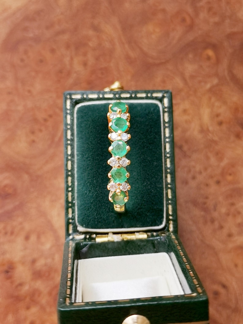 May include: A gold ring featuring a row of five round green emeralds alternating with small clear white diamonds, set in a vertical arrangement within a dark green velvet-lined jewelry box with a gold-trimmed rectangular frame.