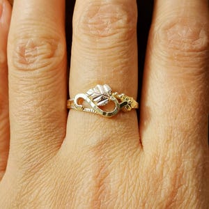 May include: A gold ring with an infinity symbol design and a leaf detail. The ring is worn on a person's finger.