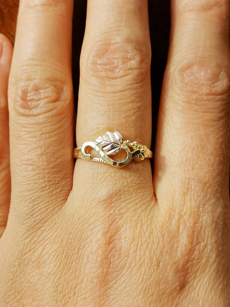 May include: A gold ring with an infinity symbol design and a leaf detail. The ring is worn on a person's finger.