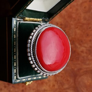 Pode incluir: Um anel de prata com uma grande gema redonda vermelha engastada em uma moldura de prata. O anel est&aacute; em uma caixa de veludo verde.