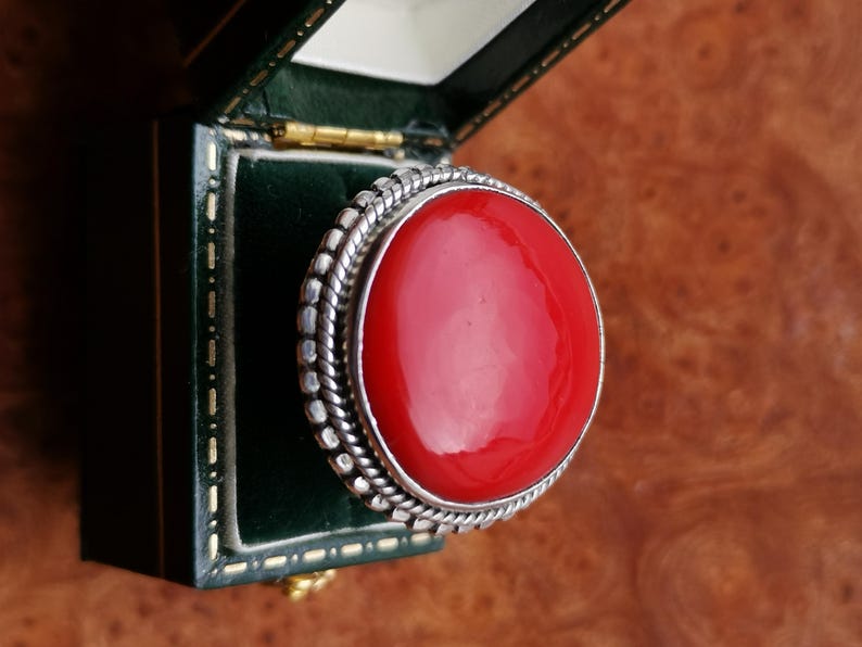 Pode incluir: Um anel de prata com uma grande gema redonda vermelha engastada em uma moldura de prata. O anel est&aacute; em uma caixa de veludo verde.