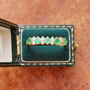 May include: A gold ring featuring a row of four round green gemstones alternating with smaller clear white stones, presented inside a dark green velvet-lined jewelry box with a gold-toned metal hinge and trim detail.
