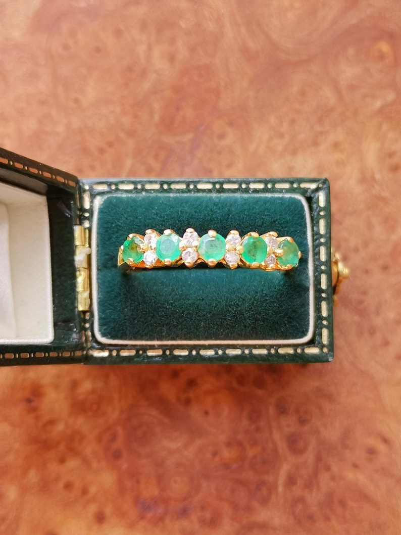 May include: A gold ring featuring a row of four round green gemstones alternating with smaller clear white stones, presented inside a dark green velvet-lined jewelry box with a gold-toned metal hinge and trim detail.