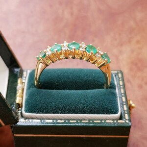 May include: A gold band ring featuring a row of five oval-cut green emeralds alternating with small clear round-cut diamonds. The ring is set in a yellow gold prong setting and is displayed inside a dark green velvet-lined jewelry box.