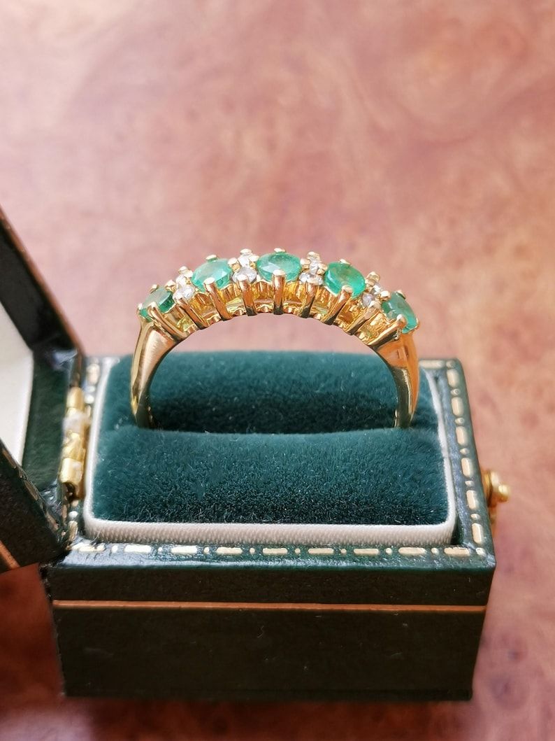 May include: A gold band ring featuring a row of five oval-cut green emeralds alternating with small clear round-cut diamonds. The ring is set in a yellow gold prong setting and is displayed inside a dark green velvet-lined jewelry box.