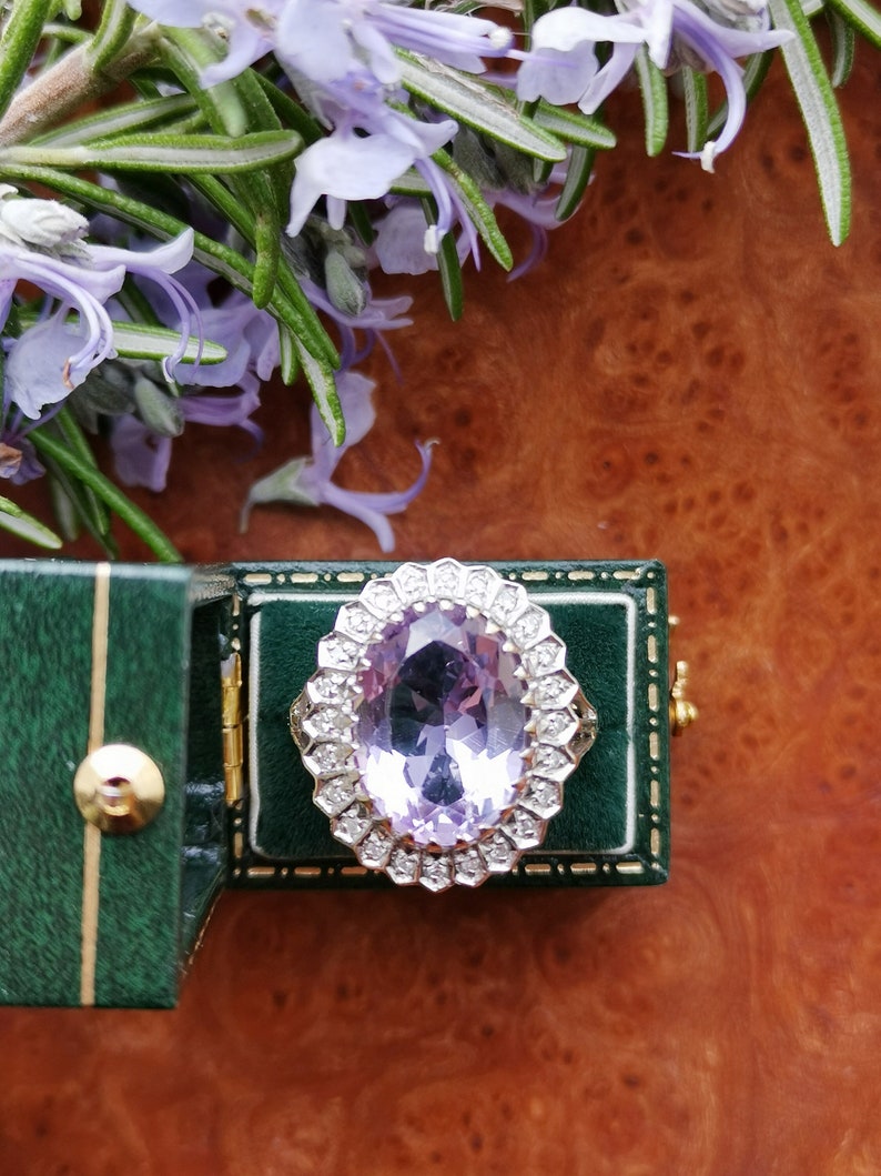 Op de afbeelding: Een ring met een grote ovale amethist-edelsteen, omringd door een halo van kleinere diamanten. De ring bevindt zich in een groene fluwelen ringdoos.