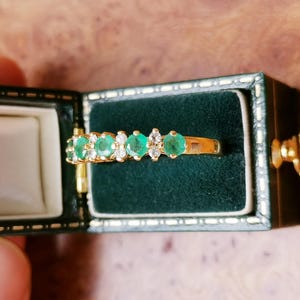 May include: A gold ring featuring a row of alternating round green emeralds and clear white diamonds set in a prong setting, displayed inside a rectangular dark green velvet-lined jewelry box with gold-toned trim and a metal latch closure.