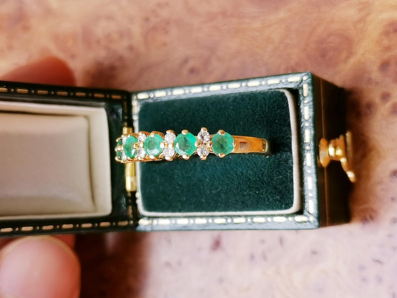 May include: A gold ring featuring a row of alternating round green emeralds and clear white diamonds set in a prong setting, displayed inside a rectangular dark green velvet-lined jewelry box with gold-toned trim and a metal latch closure.