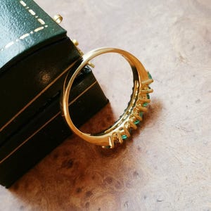 May include: A gold ring featuring a row of small, round, vibrant green gemstones set along the band. The ring is positioned at an angle, showcasing the polished metal finish and the consistent size of the green stones embedded in the setting.