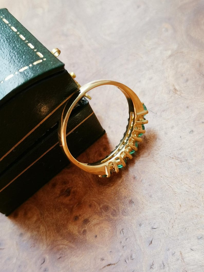 May include: A gold ring featuring a row of small, round, vibrant green gemstones set along the band. The ring is positioned at an angle, showcasing the polished metal finish and the consistent size of the green stones embedded in the setting.