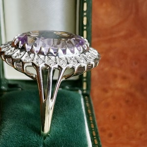 Op de afbeelding: Een zilveren ring met een grote, ovale amethist-edelsteen omringd door een halo van kleinere diamanten. De ring heeft een delicaat, ingewikkeld ontwerp en is gezet op een zilveren band.