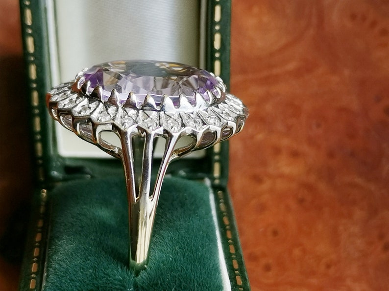 Op de afbeelding: Een zilveren ring met een grote, ovale amethist-edelsteen omringd door een halo van kleinere diamanten. De ring heeft een delicaat, ingewikkeld ontwerp en is gezet op een zilveren band.