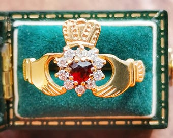 May include: A gold-colored Claddagh ring with a central red gemstone surrounded by smaller white gemstones. The ring features a crown, hands, and a heart design, set against a green velvet background.
