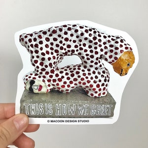 Pode incluir: Escultura de uma pessoa com bolinhas brancas e vermelhas, cabeça marrom, deitada de lado. A escultura está sobre uma base cinza com o texto "This is how we greet" e "MACOON DESIGN STUDIO".