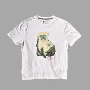 Pode incluir: Camiseta branca com um gráfico de dois macacos, um segurando o outro. Os macacos têm pelo branco, rostos azuis e detalhes verdes. O texto "Maison Design" está impresso abaixo dos macacos.