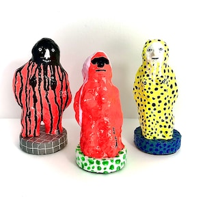 Pode incluir: Três estatuetas de cerâmica pintadas à mão. A estatueta à esquerda é vermelha e preta com um padrão listrado. A estatueta do meio é vermelha e tem um rosto branco com óculos de sol. A estatueta à direita é amarela com bolinhas pretas e um rosto branco.