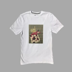 Pode incluir: Camiseta branca com uma estampa gráfica de um design abstrato vermelho e branco sobre um fundo marrom.