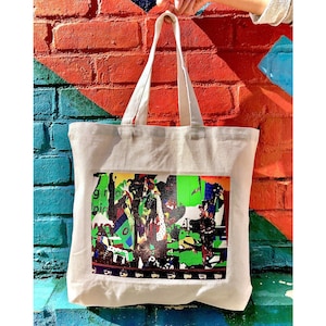 Pode incluir: Uma bolsa tote de lona branca com um design abstrato colorido impresso na frente. O design apresenta uma variedade de formas e cores, incluindo verde, vermelho, amarelo e azul.