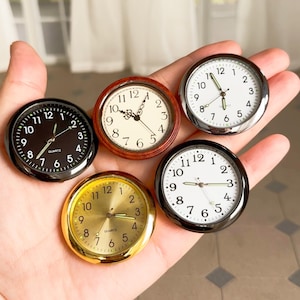 Puede incluir: Una colección de cinco pequeñas esferas de reloj de cuarzo redondas en varios estilos. Las esferas tienen esferas blancas o doradas con números y manecillas negros o dorados. Los biseles son negros, plateados, dorados y de madera.