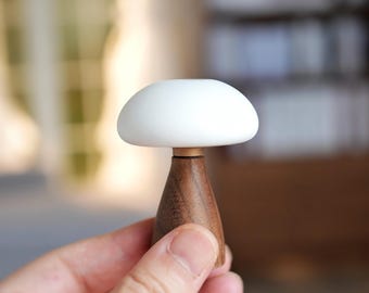 May include: A mushroom-shaped decorative object with a white, rounded top and a dark brown, conical wooden base. The base is connected to the top with a thin, light-colored ring. The object is held in a person's hand.