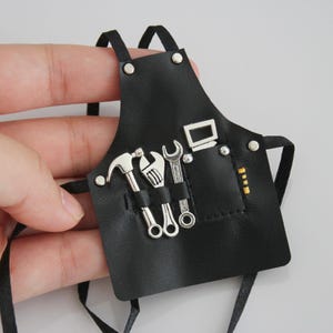 Peut inclure: Un petit tablier en cuir noir avec des décorations d'outils argentées. Le tablier comprend un marteau, une clé et d'autres outils. Il possède des sangles noires et des rivets argentés. Conçu pour une poupée ou un projet artisanal.