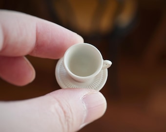 May include: A miniature white teacup and saucer set. The teacup has a small handle and sits on a matching saucer with a decorative edge. The set is made of porcelain or a similar material.