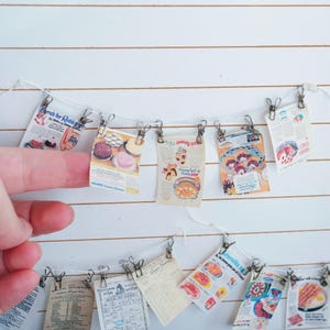 May include: A string of miniature vintage food advertisements clipped to a string with small metal clips. The ads feature colorful illustrations of various dishes, including cakes, pies, and prepared meals. The background is a white surface with horizontal lines.
