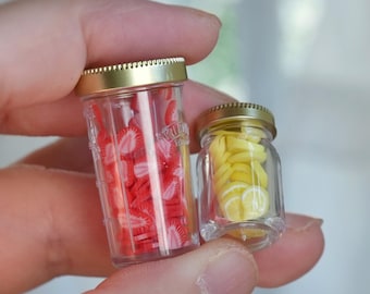 May include: Two miniature glass jars with gold lids. One jar is filled with red strawberry slices, and the other with yellow lemon slices. The jars are held in a hand.