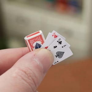 Op de afbeelding: Miniatuur Bicycle speelkaarten, met een rood en wit doosje. De getoonde kaarten zijn een schoppen aas, een harten aas en een klaveren zes. De kaarten worden in een hand gehouden.