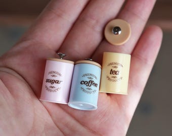 以下が含まれることがあります： 「砂糖」、「コーヒー」、「紅茶」と書かれたミニチュアのピンク、青、黄色のキャニスター。各キャニスターには木製の蓋があり、側面に「Premium Products」の文字が印刷されています。小さなベージュ色の蓋も確認できます。