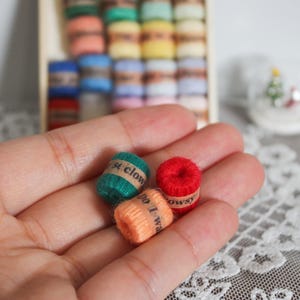 May include: A hand holding three miniature spools of thread in green, red, and orange. Each spool has a label with text. A box of additional spools in various colours is in the background, along with a small snow globe.