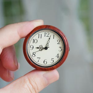 May include: A miniature round clock with a wooden frame is held. The clock face is cream with black numerals and hands. The time is approximately 1:45. The frame is a deep reddish-brown. The clock is likely a decorative item.