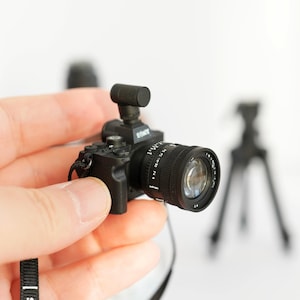 May include: A miniature black camera with a lens, flash, and strap is held in a hand. The camera has the brand name "SONY" printed on it. A small tripod is visible in the background.