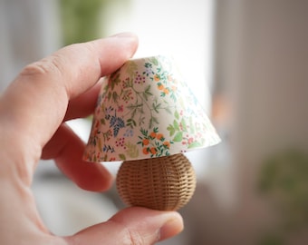 May include: Miniature lamp with a woven rattan base and a floral patterned lampshade. The lampshade features a cream background with a colorful print of flowers and foliage. The lamp is held in a hand.