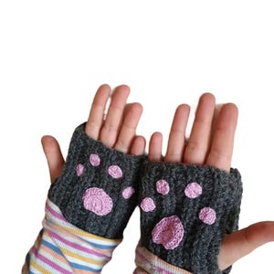 Könnte beinhalten: Ein Paar graue Fingerlose Handschuhe mit rosa Pfotenabdruck-Designs. Die Handschuhe sind gestrickt und haben eine strukturierte Oberfläche.